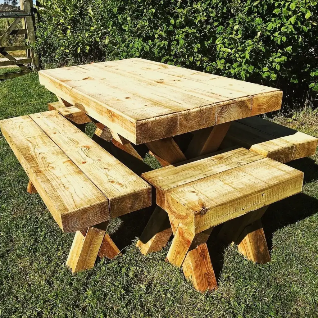 Chunky rough-sawn timber table with four thick benches on crossed legs, standing on grass