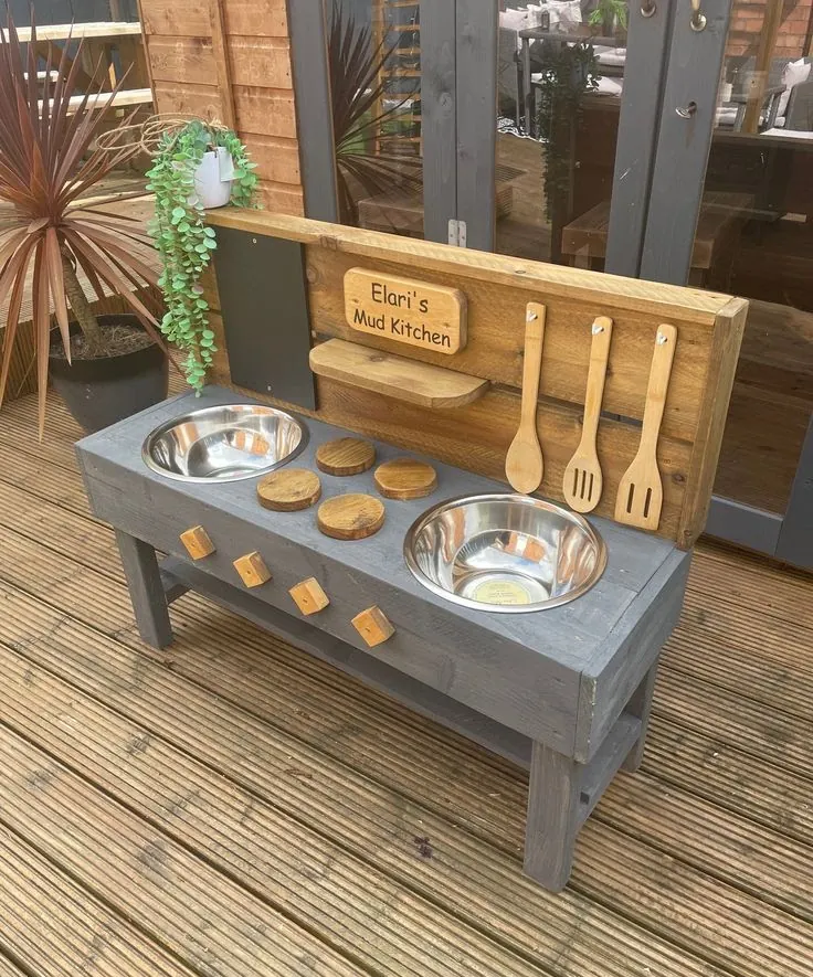 Grey mud kitchen with two bowl sinks, timber hobs and knobs, stained back panel and a name plaque