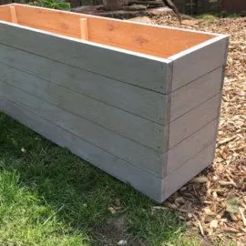 Long rectangular trough planter painted grey, set on grass beside bark chips