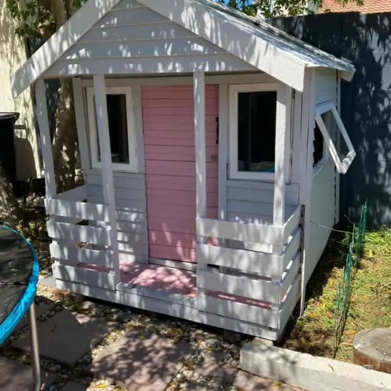 Pale grey Wendy house with a pink door, white trim, front veranda and push-out side window