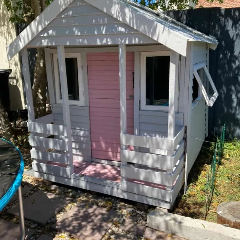 Pale grey Wendy house with a pink door, white trim, front veranda and push-out side window