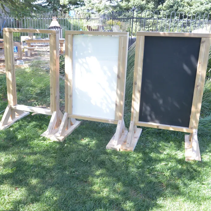Three freestanding outdoor timber easels: a clear panel, a whiteboard and a chalkboard on a lawn