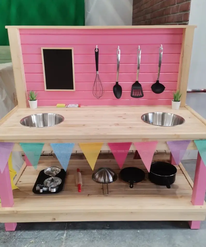 Pine mud kitchen with a pink back panel and legs, two bowl sinks, chalkboard, utensils and bunting