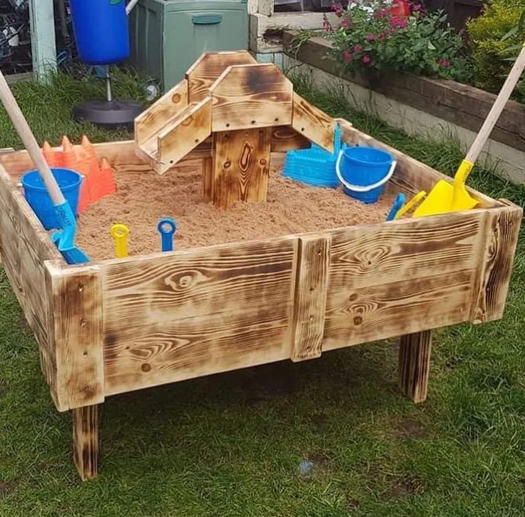 Raised sandpit on legs with a scorched wood finish and a small wooden castle in the middle