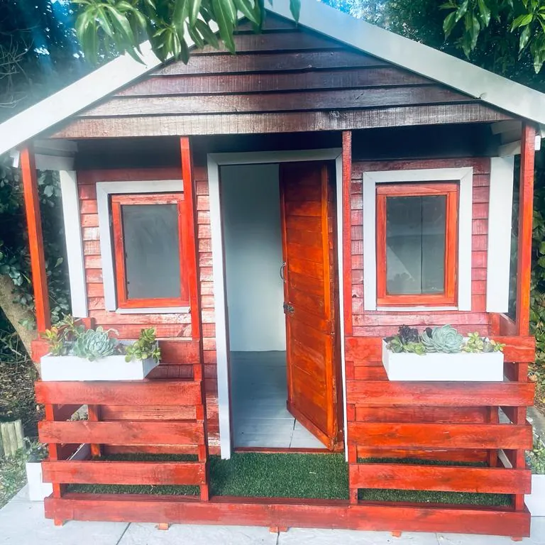 Red-stained Wendy house with white trim, open door, veranda railing and succulent window boxes