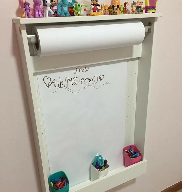 White wall-mounted paper roll easel with a top shelf for toys and pen holders on the ledge